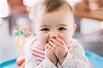 A baby girl with her hands covering her mouth, looking at the camera.