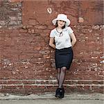 Pretty young brunette woman in a white hat, blouse and black skirt, posing outdoor in old vintage brown brick wall background. Girl is leaning against the wall. Space for text.
