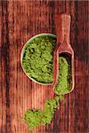 Green food supplement. Wheatgrass ground on brown wooden scoop and round bowl on brown wooden background, top view. Healthy natural detox.