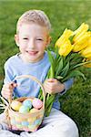 little smiling boy holding basket with colorful easter eggs and bouquet of yellow tulips after egg hunt in the park sitting on grass enjoying spring time