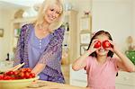 Portrait playful granddaughter covering eyes with tomatoes in kitchen