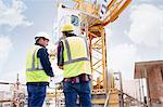 Construction worker and engineer reviewing blueprints below crane at construction site