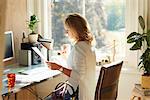 Woman drinking coffee and reading paperwork at desk in sunny home office