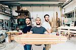 Friends standing in carpentry workshop holding skateboard looking at camera