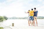 Three young adults standing on jetty, taking self portrait, using smartphone, rear view