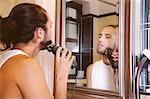 Young man looking in mirror, shaving with electric razor