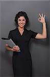 Beautiful businesswoman showing okay sign and smiling for the camera. Lady with black hair holding documents in her hand over grey.