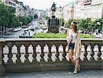 Trendy hippie woman tourist in jeans shorts, knitted shawl and white blouse standing on Wenceslas Square in Prague. In the background Saint Wenceslas statue in Prague. Tourism travel concept.