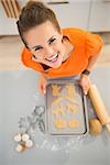 Funny modern housewife holding tray of uncooked Halloween biscuits in kitchen. Halloween treats ready to go into oven. Traditional autumn holiday. Upper view