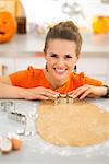 Cheerful modern woman cutting out Halloween cookies with pastry cutter from fresh rolled pastry in decorated kitchen. Traditional autumn holiday
