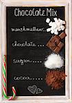 Hot chocolate mix on old chalk blackboard. Sweet marshmallow, chocolate bars, sugar, cocoa powder and mint candy cane for cooking winter holiday drink.