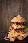 Big delicious cheeseburger stacked high with a bacon, beef patty, coleslaw, cheese, lettuce, red onion and tomato on sesame seed bun served old knife on wooden cutting board.