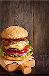 Big delicious cheeseburger stacked high with a bacon, beef patty, coleslaw, cheese, lettuce, red onion and tomato on sesame seed bun served  on wooden cutting board.