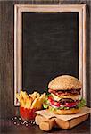 Big delicious cheeseburger stacked high with a bacon, beef patty, cheese, lettuce, red onion and tomato on sesame seed bun served  on wooden cutting board and fried potatoes with sauce.