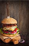 Big delicious cheeseburger stacked high with a bacon, beef patty, coleslaw, cheese, lettuce, red onion and tomato on sesame seed bun served old knife on wooden cutting board. Rustic style.