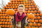 Portrait of smiling young woman holding pumpkins in front of big pyramid of pumpkins in autumn outdoors