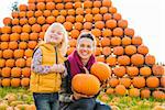 A beautiful smiling woman and cute little girl holding pumpkins in front of big pile of orange pumpkins in autumn outdoors