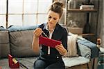 Wealthy brown-haired woman in elegant clothing sitting on couch and enjoying unpacking new gold jewelry from red box in stylish loft apartment. Luxurious life concept