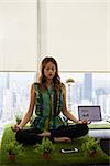 Young hispanic woman in office, sitting on desk covered with grass and plants. The businesswoman does zen and yoga meditation in lotus position. Full length, front view
