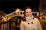 Happy elegant young woman in white coat standing in front of Rialto Bridge in the evening. She having Christmas time trip and enjoying stunning views of Venice, Italy