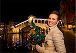 Smiling elegant young woman in white coat holding Christmas tree while standing in front of Rialto Bridge in the evening. She having Christmas time trip and enjoying stunning views of Venice, Italy