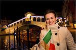 Portrait of happy elegant young woman showing Italian flag while standing in front of Rialto Bridge in the evening. She having Christmas time trip and enjoying stunning views of Venice, Italy