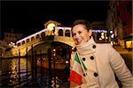 Happy elegant young woman holding Italian flag and looking on something while standing in front of Rialto Bridge in evening. She having Christmas time trip and enjoying stunning views of Venice, Italy