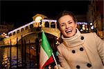 Portrait of happy elegant young woman showing Italian flag while standing in front of Rialto Bridge in the evening. She having Christmas time trip and enjoying stunning views of Venice, Italy