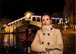 Pensive elegant young woman in white coat standing in front of Rialto Bridge in the evening. She having Christmas time trip and enjoying stunning views of Venice, Italy