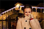 Elegant young woman in white coat holding collar of coat while standing in front of Rialto Bridge in the evening. She having Christmas time trip and enjoying stunning views of Venice, Italy