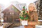Christmas season in relaxed style of contemporary countryside living. Young woman in furry hat holding Christmas tree and looking into distance while standing in the front of a cosy mountain house.