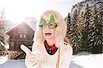 Going wild for Christmas season in a secluded spot in the countryside. Smiling young woman in funny Christmas tree glasses pointing on something in the front of a cosy mountain house.