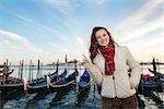 Sunset brings to life irresistible magic of Venice - the unique Italian city. Happy young woman traveler standing on embankment in the front of the line of gondolas and pointing on copy space