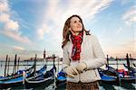 Sunset brings to life irresistible magic of Venice - the unique Italian city. Dreamy young woman traveler standing on embankment in the front of the line of gondolas and looking into distance