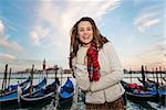 Sunset brings to life irresistible magic of Venice - the unique Italian city. Portrait of happy young woman traveler standing on embankment in the front of the line of gondolas