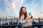 Sunset brings to life irresistible magic of Venice the unique Italian city. Young woman traveler with Italian flag standing on embankment in the front of the line of gondolas and looking into distance