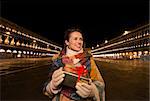 Excitement of Christmas time and allure of long-time favourite Venice, Italy. Happy woman holding Christmas gift box while standing on Piazza San Marco in the evening