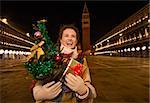 Excitement of Christmas time and allure of long-time favourite Venice, Italy. Smiling woman holding Christmas tree and present box while standing on Piazza San Marco in the evening