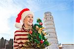 Young, itching from energy and searching for excitement. I'm going to Christmas trip to Italy. It is no-brainer. Woman in Santa hat with Christmas tree looking on something near Leaning Tour of Pisa.