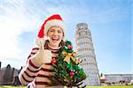 Young, itching from energy and searching for excitement. I'm going to Christmas trip to Italy. It is a no-brainer. Woman in Santa hat with Christmas tree showing thumbs up near Leaning Tour of Pisa