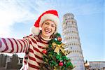 Young, itching from energy and searching for excitement. I'm going to Christmas trip to Italy. It is a no-brainer. Woman in Santa hat with Christmas tree taking selfie near Leaning Tour of Pisa