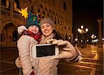 Holiday family trip to Venice, Italy can change the whole Christmas experience. Happy mother and child in funny Christmas hat taking selfie on Piazza San Marco in the evening