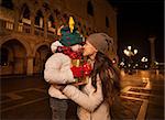 Holiday family trip to Venice, Italy can change the whole Christmas experience. Happy mother hugging child with Christmas gift box while standing on Piazza San Marco in the evening