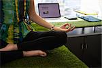 Young hispanic woman in office, sitting on desk covered with grass and plants. She meditates yoga in lotus position. Cropped view of hands and feet