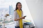 Young multiethnic Chinese Hispanic woman leaning on table in a modern office building, with a beautiful sight of the city in background. She holds a mobile phone and smiles at camera