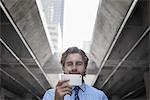 A man on a city street, holding a mobile phone taking a selfie, with an urban walkway and tall buildings in the background.