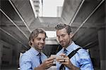 Two men in shirts and ties taking a selfie with a smart phone, with an urban walkway and tall buildings in the background.