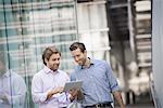 Two men standing outside a large building, one holding a digital tablet.