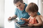 A man and a young child sitting looking at a digital tablet and touching the screen.