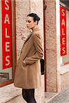 cute pretty brunette woman near a window-shop looking for winter sale . she is wearing a coat and a bag